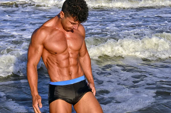 Strong bodybuilder with six pack.Fitness trainer with perfect abs, shoulders,biceps, triceps,chest, flexing his muscles on the beach with sea waves on the background, training in vacation