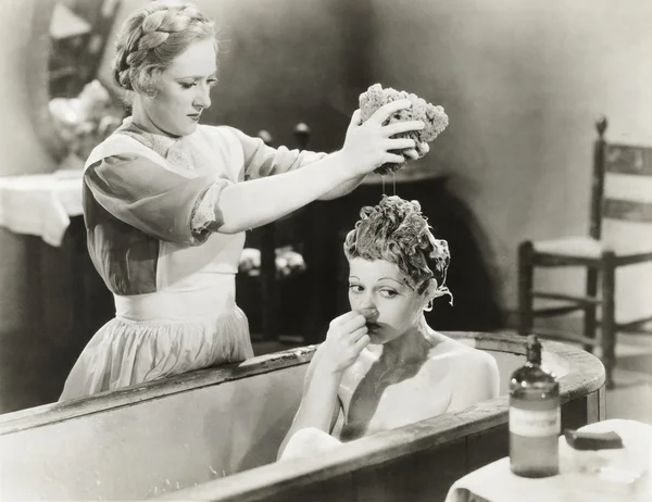 Maid squeezing sponge on woman in bathtub
