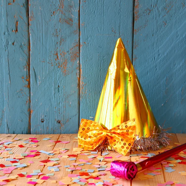 Party hat next to pink party whistle on wooden table with colorful confetti. vintage filtered image
