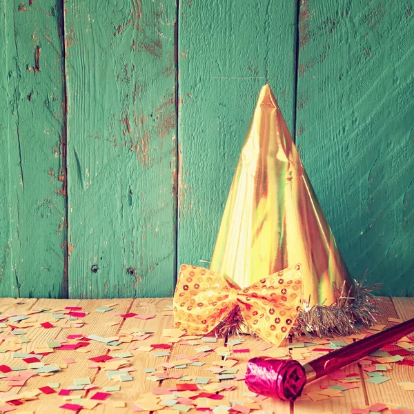 Party hat next to pink party whistle on wooden table with colorful confetti. vintage filtered image