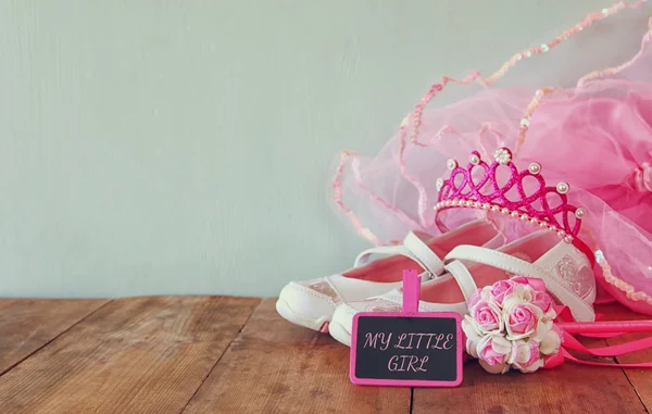 Small girls party outfit: white shoes, crown and wand flowers next to small chalkboard with phrase MY LITTLE GIRL: on wooden table. bridesmaid or fairy costume. glitter overlay