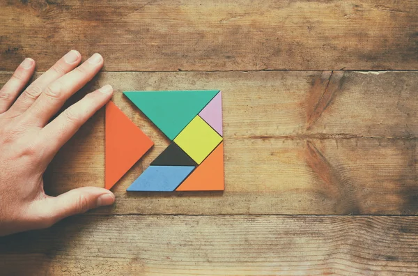 Man\'s hand holding a missing piece in a square tangram puzzle, over wooden table.