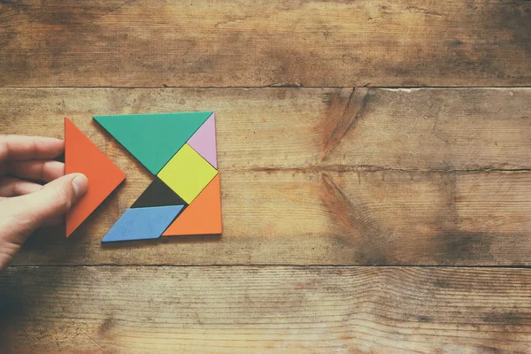 Man\'s hand holding a missing piece in a square tangram puzzle, over wooden table.