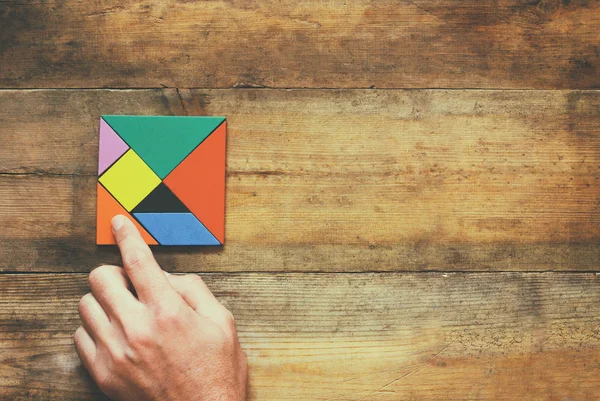 Man\'s hand holding a missing piece in a square tangram puzzle, over wooden table.