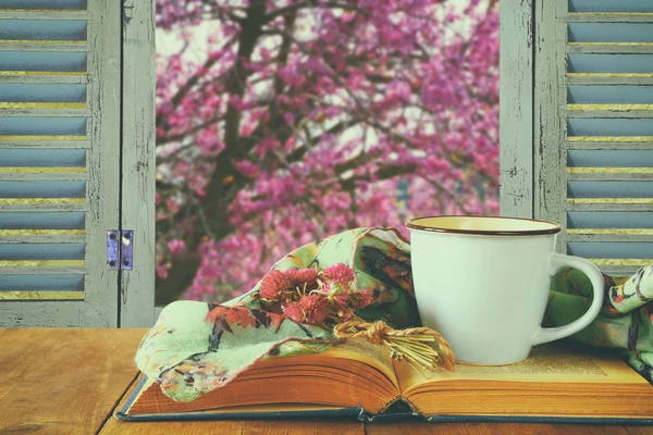Romantic scene of cup of coffee next to old book in front of countryside view outside of the old rustic window. vintage filtered image