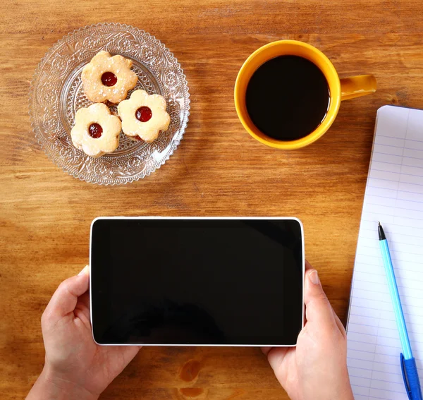 Top view of woman hands holding tablet device with empty screen, with cookies and coffee cup. image is retro filtered