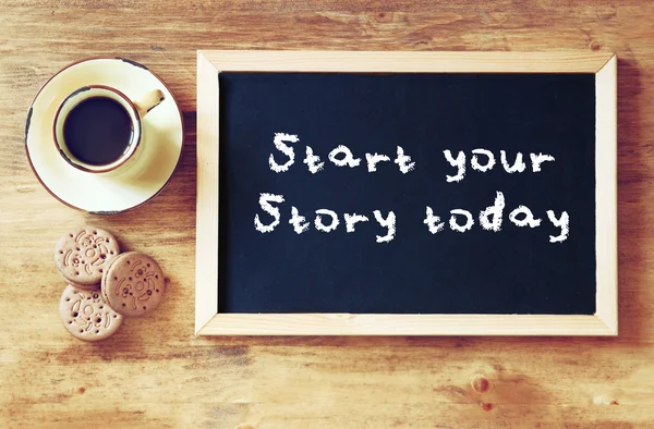 Top view of blackboard with the phrase start your story today next to cup of black coffee and cookies