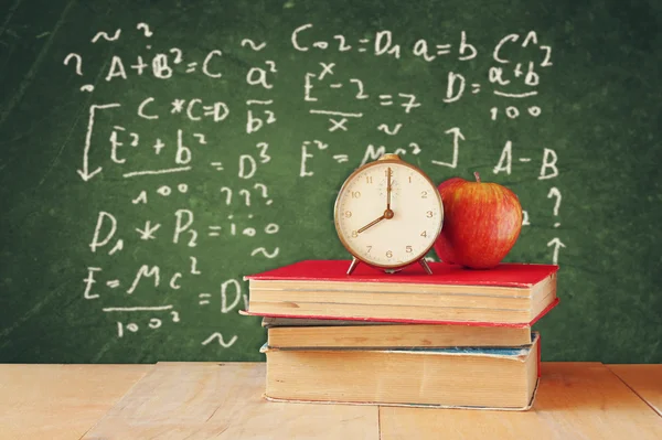 Image of school books on wooden desk, apple and vintage clock over green background with formulas. education concept