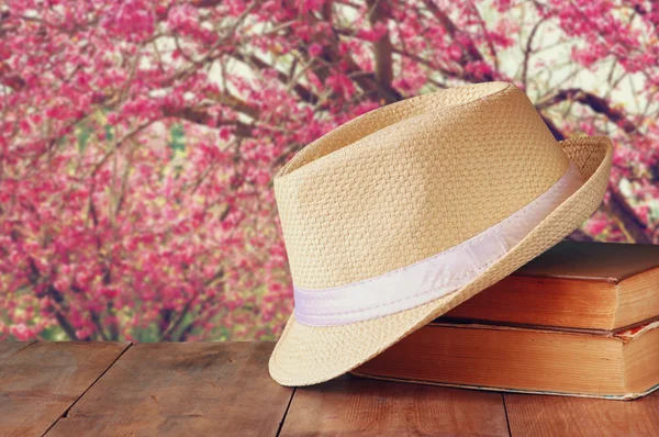 Fedora hat and stack of books over wooden table and cherry blossom tree landscape background. relaxation or vacation concept
