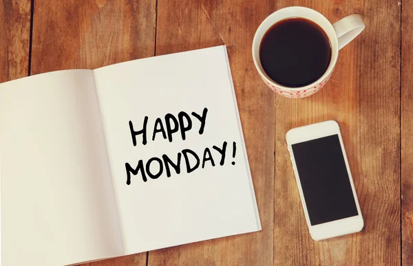 Notebook with the phrase happy monday written on it, coffee cup and smart phone. filtered image