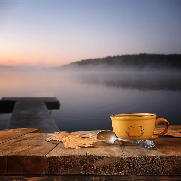 Front image of coffee cup over wooden table in front of calm foggy lake view at sunset.