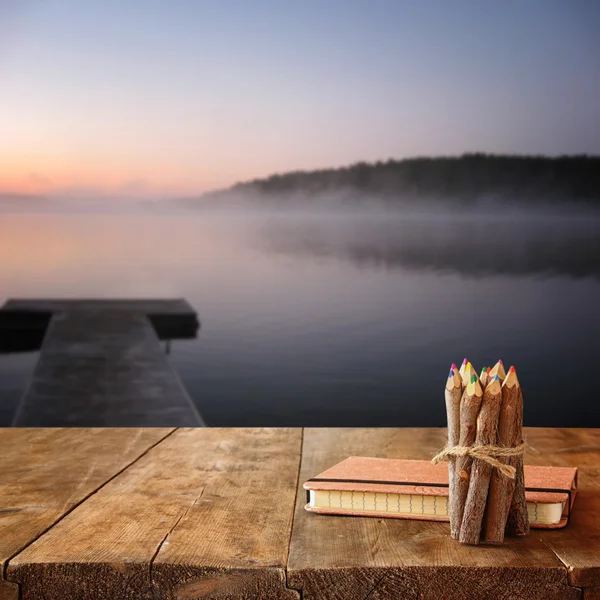 Vintage notebook and stack of wooden colorful pencils on wooden texture table in front of calm foggy lake view at sunset.