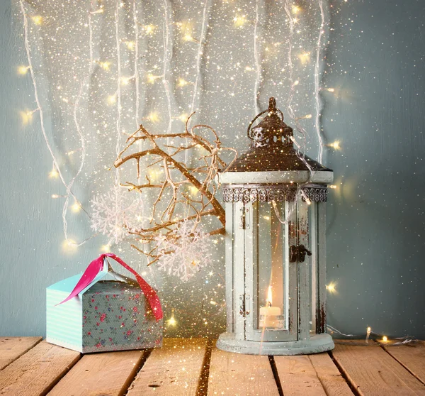 White wooden vintage lantern with burning candle christmas gifts and tree branches on wooden table. retro filtered image with glitter overlay.