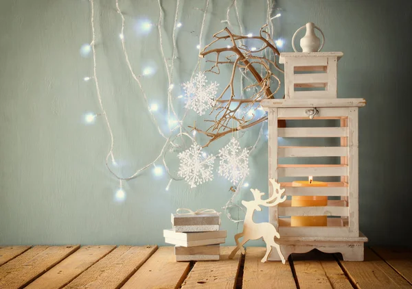 White wooden vintage lantern with burning candle, wooden deer, christmas gifts and tree branches on wooden table. retro filtered image.