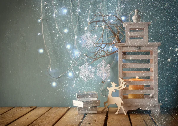 White wooden vintage lantern with burning candle, wooden deer, christmas gifts and tree branches on wooden table. retro filtered image with glitter overlay.