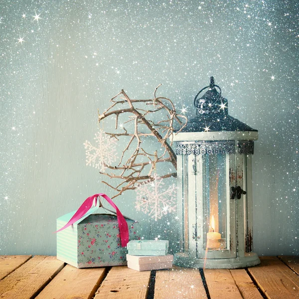 White wooden vintage lantern with burning candle christmas gifts and tree branches on wooden table. retro filtered image with glitter overlay.