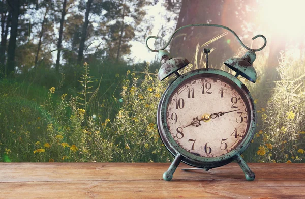 Image of vintage alarm clock on wooden table in front of rustic country side landscape background. retro filtered.