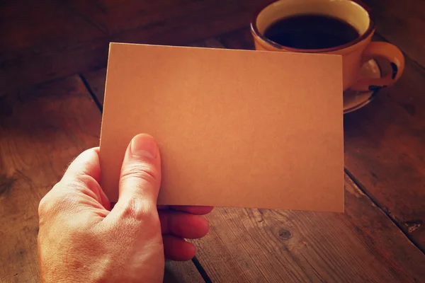 Male hands holding brown empty card over wooden table background and cup of coffee. retro style image, low key and warm tones.