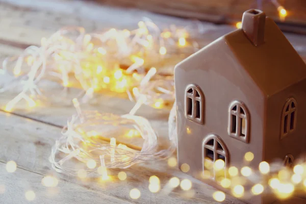 Decorative house next to gold garland lights on wooden background. copy space.