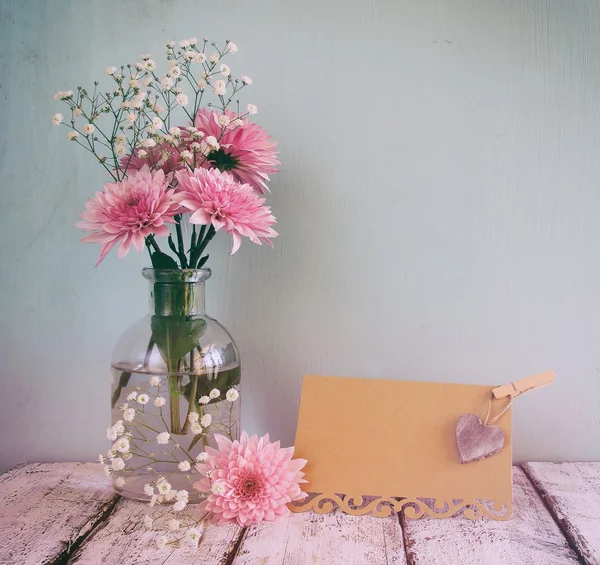 Fresh white and pink flowers, heart next to vintage empty card over wooden table. vintage filtered and toned image