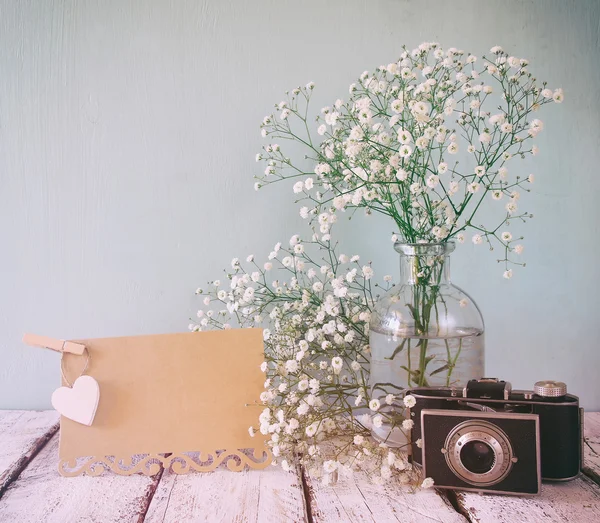 Fresh white flowers, heart next to vintage empty card and old camera over wooden table.