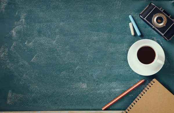 Top view image of cup of coffee, notebook and vintage camera over blackboard background