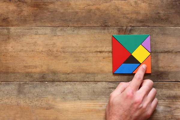 Man\'s hand holding a missing piece in a square tangram puzzle, over wooden table.