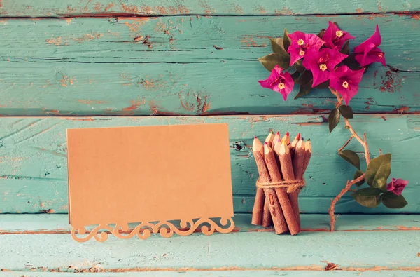 Purple bougainvillea flowers, colorful wooden pencils next to vintage empty card over wooden table. vintage filtered