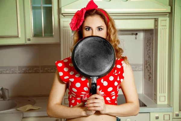 Girl in pin up style posing in the kitchen with frying pan in ha