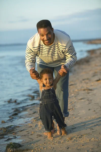 African father helping toddler walk on beach