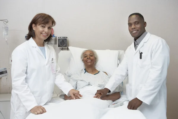 African female and male doctors holding hands with senior patient in hospital bed