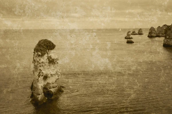 Lonely rock. Beautiful stone formations, arches and caves at Dona Ana Beach and sailing boats on horizon (Lagos, Algarve coast, Portugal) in the evening light. Retro aged sepia photo with scratches.