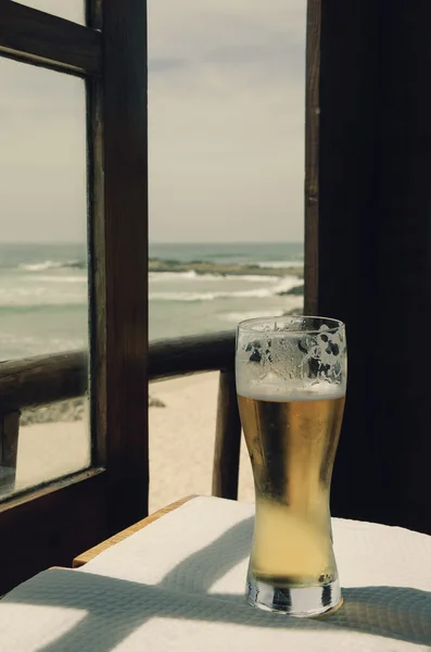 Cold beer on cafe terrace with the view on the ocean beach through the opened window. Algarve, Portugal. A game of light an shadow. Aged photo.