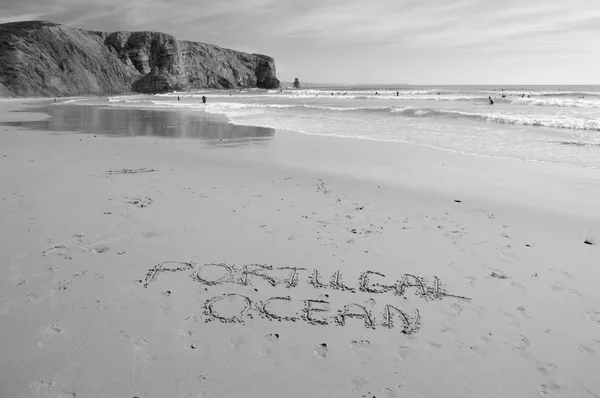 Words PORTUGAL and OCEAN are written on the scenic beach in the Algarve region of Portugal. People swimming and surfing. Aged photo. Black and white.