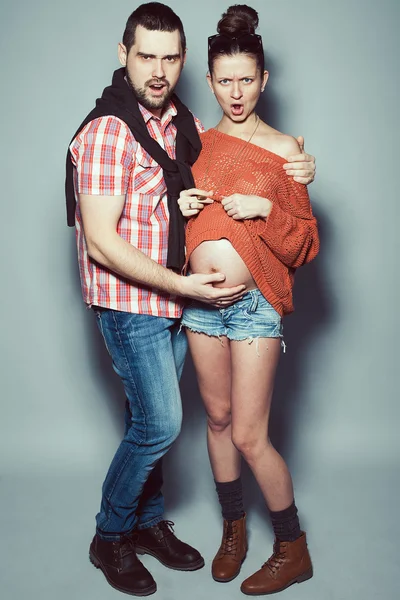 Stylish pregnancy & family concept: portrait of disturbed couple of hipsters (husband and wife) in trendy casual clothing, eyewear posing over gray background. Urban street style. Studio shot