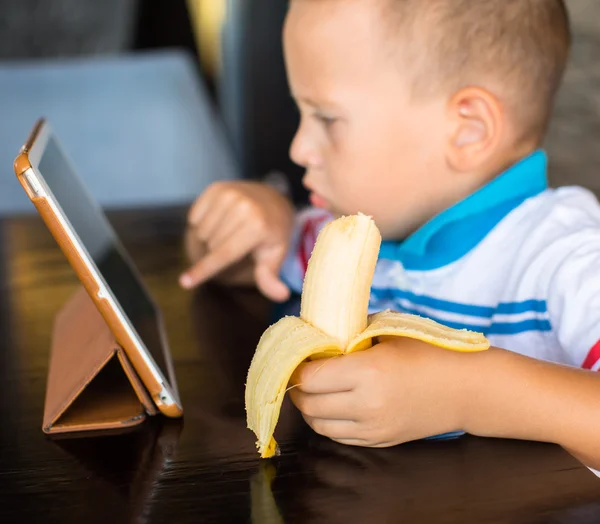 Adorable boy, eating his banana, while watching movie on tablet