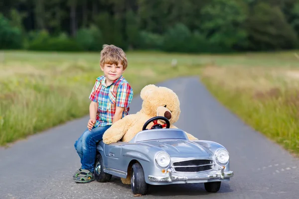 Little kid boy driving big toy car with a bear, outdoors.