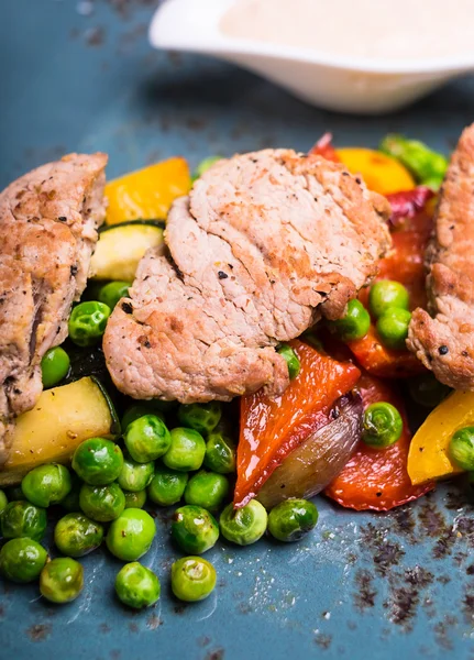 Roasted pork meat fillet chops with zucchini, onion, yellow and orange bell pepper, frozen peas on a plate with cream sauce, selective focus