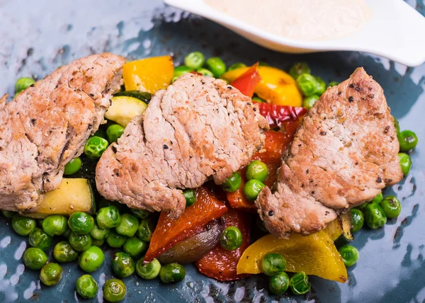 Roasted pork meat fillet chops with zucchini, onion, yellow and orange bell pepper, frozen peas on a plate with cream sauce, selective focus