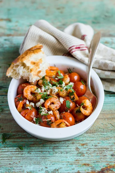 Traditional mediterranean dish with roasted cherry tomatoes, shrimps, salted greek feta cheese and fresh parsley in a bowl with homemade fresh bread on a wooden table, selective focus