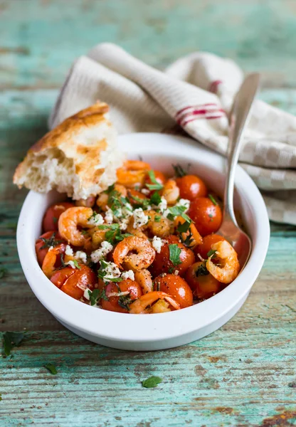 Traditional mediterranean dish with roasted cherry tomatoes, shrimps, salted greek feta cheese and fresh parsley in a bowl with homemade fresh bread on a wooden table, selective focus