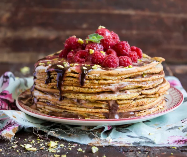 Stack of wheat golden pancakes or pancake cake with freshly picked raspberry, chopped pistachios, chocolate sauce on a dessert plate