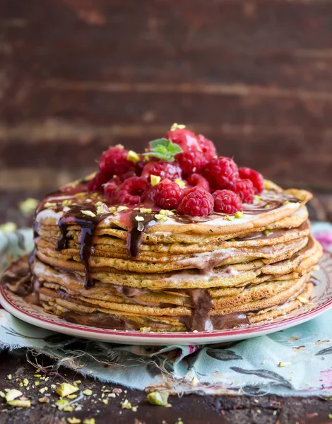 Stack of wheat golden pancakes or pancake cake with freshly picked raspberry, chopped pistachios, chocolate sauce on a dessert plate