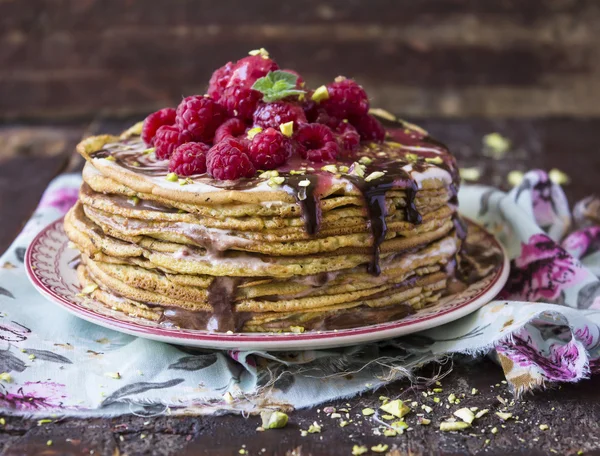 Stack of golden wheat pancakes with chocolate sauce, pistachios, fresh raspberry and mint on a dessert plate on a wooden rustic table, selective focus