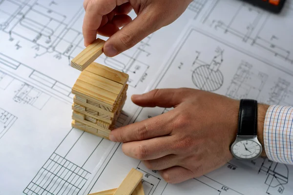 Man architect draws a plan, graph, design, geometric shapes by pencil on large sheet of paper at office desk and builds model house from wooden blocks (bars)