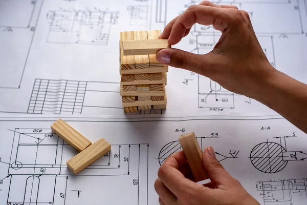 Man architect draws a plan, graph, design, geometric shapes by pencil on large sheet of paper at office desk and builds model house from wooden blocks (bars)
