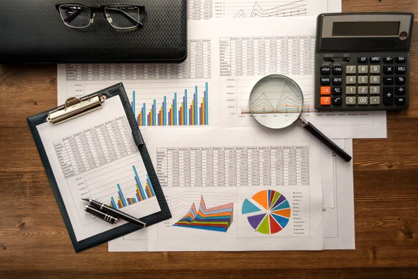 Business accessories (notebook, calculator, planchette, tablet, fountain pen, glasses) and graphics, tables, charts on white sheets on office desk. Soft focus.