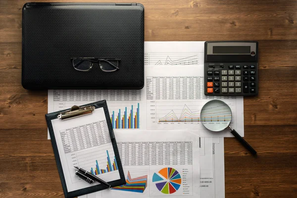 Business accessories (notebook, calculator, planchette, tablet, fountain pen, glasses) and graphics, tables, charts on white sheets on office desk. Soft focus.