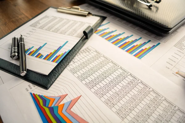 Business accessories (notebook, calculator, planchette, tablet, fountain pen, glasses) and graphics, tables, charts on white sheets on office desk. Soft focus.