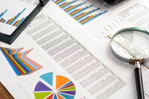 Business accessories (notebook, calculator, planchette, tablet, fountain pen, glasses) and graphics, tables, charts on white sheets on office desk. Soft focus.